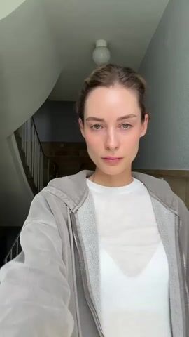 A woman stands in a tiled stairwell. She is fair-skinned and has her hair pulled back. She wears a white t-shirt, a gray zip-up hoodie, and a long, flowing, light pink skirt. A white undergarment is visible beneath her t-shirt. She puts on a pair of dark sunglasses, turns her back briefly, and then faces the camera again.  Behind her is a stairwell with white railings. A small black dolly or buggy is visible on the stairs. The walls are light gray and tiled, and there is a simple light fixture on the ceiling.
