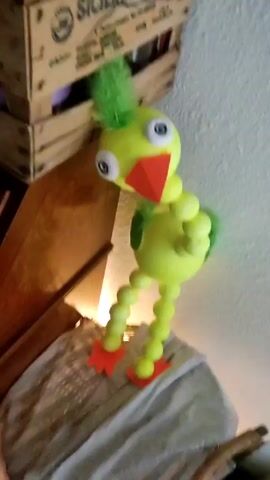 A video pans from a yellow, cartoonish chicken-like figure with a red beak to a framed silhouette of a woman holding flowers. The chicken has a green tuft of hair and legs made of small, yellow balls. The woman silhouette is black, with white and colorful accents. The objects are near a white wall. Dance music is playing in the background.