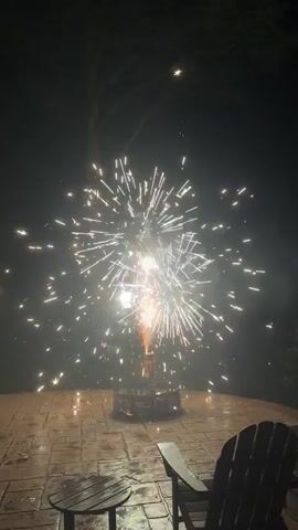 The video captures a nighttime scene with fireworks being set off in a fire pit located on a paved patio. Two chairs are visible in the foreground, one of which is an Adirondack chair. The fireworks create a bright burst of white sparks that spread outwards against the dark sky. The scene appears to be set in a backyard or outdoor recreational area.