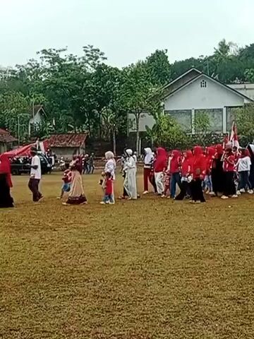 A group of people are walking across a grassy field. Many of them are wearing red and white. Some are carrying flags. In the background, there are houses and trees. The sky is overcast.