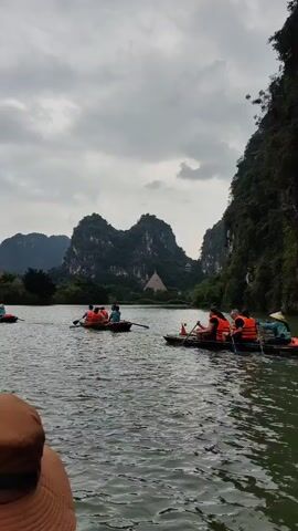 The video depicts a tranquil scene on a river or lake surrounded by lush, mountainous terrain under an overcast sky. Several small boats, each carrying a group of people wearing orange life vests, are visible on the water. These boats appear to be rowed by one or two individuals in each. The background is dominated by jagged, tree-covered mountains, providing a dramatic backdrop to the waterway. A hint of the videographer's hat is visible in the foreground, suggesting that the scene is captured from the perspective of someone in a similar boat. The overall impression is that of a leisurely tourist activity in a scenic location.