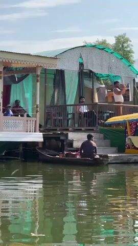 The video shows a houseboat on a body of water. The houseboat appears to be made of wood and is decorated with colorful fabric. Several people are visible on the houseboat, including a man standing on the deck and taking a picture with his phone, and another man sitting in a boat in the foreground. The water is calm and reflects the houseboat and the surrounding scenery. In the background, there are mountains and trees. The overall impression is one of a peaceful and scenic location.