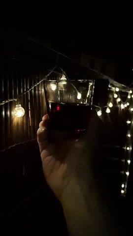 A person's hand holds up a glass filled with dark red liquid. The glass has a textured design and is illuminated by string lights. The background is dark, with some of the lights reflecting off a ribbed surface.