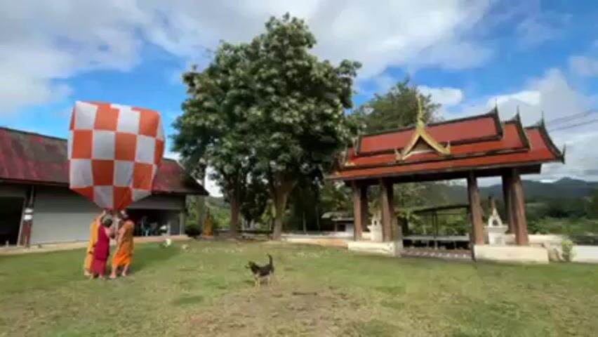 Three monks stand on a grassy field in front of a building with a red roof, holding a large, square balloon with an orange and white checkerboard pattern. A small dog stands in front of them, facing the camera. Behind the monks and to the right, there is a traditional Thai-style pavilion with a red tiled roof and white posts. A large tree is in the center of the shot. The sky is blue with scattered white clouds.