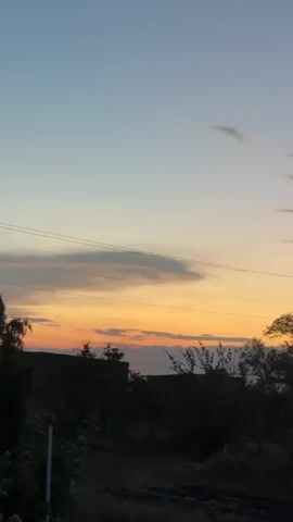 A vertical video shows a rural landscape at dusk. The sky transitions from a pale blue at the top to a warm orange and yellow near the horizon, with scattered clouds. Silhouetted against the sky are trees and what appears to be a building or structure on the left. Power lines run horizontally across the frame. The foreground is dark and indistinct, with some vegetation and what could be discarded materials on the ground. The overall impression is of a quiet, end-of-day scene in the countryside.