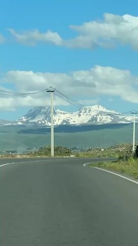 This media file captures a scenic view of a winding asphalt road with white lane markings, leading towards majestic snow-capped mountains. A solitary, tall utility pole with multiple wires stretches upwards, bisecting the frame and adding a linear element to the landscape. The mountains, dusted with snow, rise prominently against a vibrant blue sky dotted with fluffy white clouds. Below the mountains, rolling green hills are visible, interspersed with what appears to be a small village or scattered houses, suggesting a rural or semi-rural setting. The terrain in the foreground and along the roadside is rocky and covered with sparse vegetation.

The image conveys a sense of journey and natural beauty. The time of day appears to be daytime, likely late morning or afternoon, given the bright sunlight and clear skies. The weather seems pleasant, with sunshine illuminating the landscape and casting soft shadows. The overall mood is tranquil and picturesque, inviting contemplation of the vastness of nature and the open road. The presence of the road and the utility pole hint at human presence and infrastructure within this natural environment. The location is identified as Horom, Armenia, providing specific geographical context to the observed scene. There is no visible text within the media file.