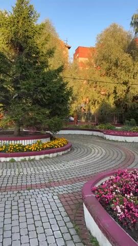 A video shows a paved walkway with pink and gray interlocking bricks forming a circular pattern. On the sides of the walkway are curved flower beds with pink and yellow flowers. There are trees and bushes surrounding the walkway, and a brick building can be seen in the background. The sun is shining, casting shadows.