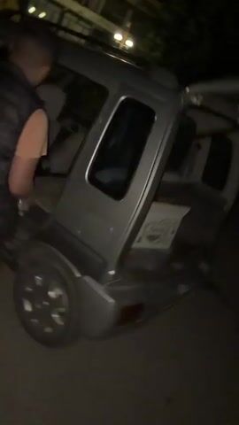 A person climbs into a silver car through the trunk. The person is wearing a dark, possibly black, patterned shirt and dark pants. The car appears to be parked at night, with some ambient light visible in the background. The video is filmed from a slightly elevated perspective, looking down at the car.