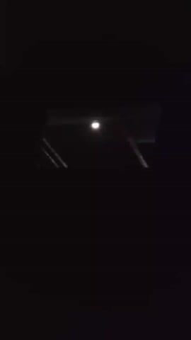 The video captures a dark, nocturnal scene, likely filmed from inside a room looking out through a window or opening. The primary subject is a bright, circular light source, possibly the moon or a distant streetlamp, suspended in the darkness. This light is positioned within a rectangular frame, suggesting it's viewed through a window with a visible sill or frame.

The surrounding environment is almost entirely black, with minimal discernible details. However, faint diagonal lines can be observed within the frame, hinting at structural elements or perhaps reflections. The overall impression is one of nighttime stillness.

The audio consists of muffled human voices speaking in Indonesian. One person can be heard saying, "bulannya yang ... kayak gini," which translates to "the moon is like this." This suggests the speakers are observing and commenting on the celestial object visible in the frame. The tone seems casual and observational, with no indication of distress or excitement.

The time of day is clearly night, as indicated by the darkness and the presence of the moon or a light source. There are no weather cues, but the darkness implies a clear night. The location context of Karangkemiri, Indonesia, doesn't provide specific visual cues within the frame itself, but it anchors the observation to a particular geographical setting. No visible text is present in the video. The emotions conveyed are subdued curiosity and quiet observation of the night sky.