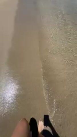 The video shows a sunny day at the beach. The camera follows a person walking barefoot in the shallow, clear water along the shoreline. The sand is wet near the water and dryer further up the beach. Small waves gently break on the shore. A small group of people are visible in the distance further up the beach. The sky is clear and blue. There is a small area of trees at the far end of the beach.