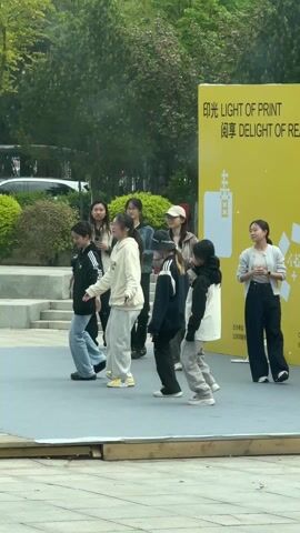 A group of about ten young women are gathered on a small stage in what appears to be an outdoor public space. Six of the women are standing on the stage facing the audience. They appear to be rehearsing a dance or performance, as they are moving in unison and slightly out of sync. The other four women are standing behind them. They seem to be observing. Behind the group is a large yellow sign with white text in both English and Chinese. The words “LIGHT OF PRINT” and “DELIGHT OF RE” are visible in English. The backdrop features trees, bushes, and a paved area. A white car is visible in the background.