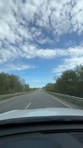 The video captures a first-person perspective from inside a vehicle traveling on a highway. The camera is pointed forward through the windshield, showing the road ahead, the sky, and the surrounding landscape. The sky is a bright blue with scattered white clouds. On either side of the highway are trees and vegetation. The road appears to be a two-lane highway with a clear lane divider. There are guardrails on the sides of the road. In the distance, other vehicles can be seen on the highway. In one brief segment, a red motorcycle is visible in the left lane, passing by the camera's vehicle. The lighting suggests it is daytime, possibly late morning or afternoon.