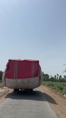 A video shows a wide, oversized load being transported on a truck on a narrow road. The load is covered with a red tarp on top and a light-colored tarp on the bottom, making it appear as a large, roundish shape. The truck is moving slowly on a paved road flanked by dirt shoulders and fields of crops. In the distance, a person on a motorcycle approaches from behind the truck. The sky is clear and blue.