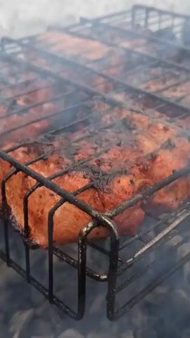 The video shows food being grilled on a barbecue rack, surrounded by a smoky haze. The food, appearing to be pieces of meat, are arranged closely together within the rack. The rack's metal bars, black in color, create a grid-like pattern. A fair amount of charring is visible on the meat, giving it a darkened color in places. The smoke obscures much of the background, leaving the viewer with little information about the environment beyond the grill.