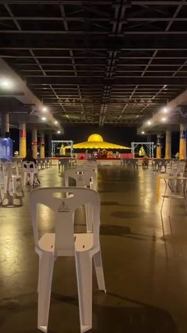 The video shows an outdoor gathering area at night, likely for a religious ceremony. Rows of white plastic chairs are set up facing a raised platform. In the background, a large, glowing golden dome structure is prominently displayed. On the platform, two individuals in saffron robes, presumably monks, are seated, facing each other. The area is illuminated by overhead lights and spotlights, casting a warm glow on the scene. Several other people are visible sitting in chairs further back, though their faces are not clear. Large golden bells hang from structures on either side of the platform. The overall atmosphere appears calm and reverent.