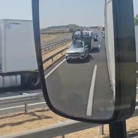 A video taken from a vehicle shows a silver Mercedes-Benz SUV towing a small white caravan on a highway. Two bicycles are mounted on a roof rack on top of the SUV. The caravan has tinted windows and a light-colored stripe along its side. The SUV is in the right lane of a multi-lane highway. In the distance, there are other vehicles, including trucks and cars, and the highway appears to have a moderate amount of traffic. The surrounding environment is dry and arid, with sparse vegetation and some trees visible on the side of the road. The sky is clear and blue, suggesting a sunny day. The video is taken from the perspective of someone inside a larger vehicle, possibly a truck, looking out through a side mirror and a side window. The reflection in the side mirror provides a distorted but clear view of the SUV and caravan. The camera is slightly shaky, indicating movement.