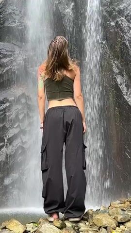 A person stands with their back to the camera, facing a large waterfall. The individual has long, light brown hair, and is wearing a dark olive green sleeveless crop top that exposes their lower back and a small portion of their midriff. They are also wearing loose-fitting black cargo pants. A tattoo is visible on their upper left arm and shoulder area, appearing to be colorful. The person stands on a rocky surface at the base of the waterfall. The waterfall cascades down a dark, textured rock face covered in some patches of greenery. The person slightly sways their hips and briefly turns their head from side to side as they observe the waterfall. The sound of rushing water is prominent.