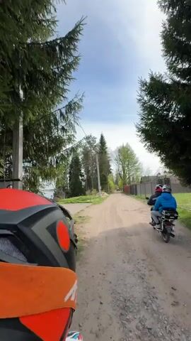 The video shows a first-person view from behind a jet ski as it travels down a dirt road. The jet ski is black with red and orange accents. On the left, there's a green tree and a glimpse of a body of water, possibly a river or a lake. The water extends into the distance. Beyond the road, there are several green trees of varying sizes. There's also a wooden pier and a small building visible near the waterfront. The sky is partly cloudy. On the right side of the road, there's a fence and a utility pole. The overall scene is a rural or waterfront landscape.