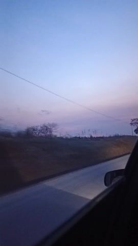 A car travels on a road during twilight. The sky is a mix of blue and purple hues. On the side of the road is a grassy hill, dotted with trees. Power lines stretch across the sky. The video is shot from inside the car, with part of the car's interior visible.