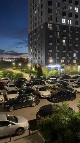 The video begins with a low-angle shot looking up at a tall, modern apartment building at dusk or early night. The building has many windows, some of which are illuminated. In the foreground, a densely packed parking lot is visible, filled with numerous cars of various colors and models. The lighting in the parking lot appears to be from street lamps.

As the video progresses, the camera pans to the right, revealing a broader view of the urban landscape. The sky is dark blue with some clouds, and a bright full moon is visible, partially obscured by clouds. More buildings, appearing to be residential, are seen in the distance, with lights on in some of their windows. Several streetlights are also visible, illuminating parts of the parking lot and the surrounding area. The overall impression is a typical residential area at night, with a high-rise building dominating the immediate foreground. There are no visible people in the video.