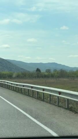 The video shows a road bordered by a guardrail in a mountainous area. There is a gray dump truck driving ahead. Another dump truck is visible in the distance. The sky is blue with some clouds.