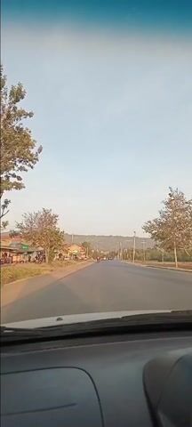 The video captures a forward-facing view from inside a car driving along a road in Machakos, Kenya. The sky is clear and bright, suggesting daytime conditions, possibly morning or late afternoon.

On the left side of the road, buildings, some appearing to be shops, and trees line the street. Utility poles and power lines are visible overhead on both sides of the road. A hilly landscape can be seen in the distance.

At the beginning of the video, a man in a dark jacket and cap, riding a motorcycle (locally known as a boda-boda), pulls out from the left side onto the main road in front of the recording vehicle. He proceeds to drive ahead.

As the car continues, other vehicles enter the frame. These include a silver minivan with the license plate "KBY 335E", a white pickup truck marked with "KBP 315E" and yellow and red reflective tape, and various other cars. Further down the road, a purple/pink matatu (minibus, a common form of public transport in Kenya) is also seen. The scene depicts typical road traffic and the general street environment of a Kenyan town or rural area.