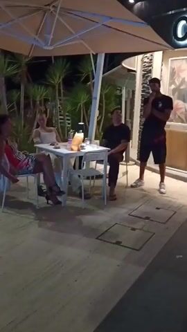 This video shows an outdoor seating area at night, likely a restaurant or lounge, with the sign "LIDO COSTA EST" prominently displayed. Several people are seated at tables, some appearing to be dining or socializing. A man is standing and speaking, possibly entertaining or addressing the crowd. Another man is seated and gestures with his arm raised. The setting appears to be a lively evening, possibly a vacation spot given the palm trees and outdoor ambiance. The lighting suggests it's after dark, and the atmosphere seems relaxed and enjoyable. No specific weather conditions are discernible, but the outdoor setting implies pleasant weather.
