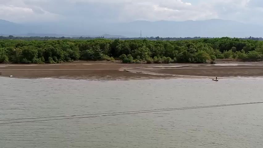 The video shows a wide, calm body of water with a muddy shoreline. A dense forest of green trees borders the shoreline, with mountains in the background under a cloudy sky. There are two small figures visible on the muddy bank in the distance on the left, and one person standing in the water on a small floating device on the right. Two parallel wires are visible spanning across the water in the foreground. The video is static and there is no movement or action beyond the stillness of the scene.