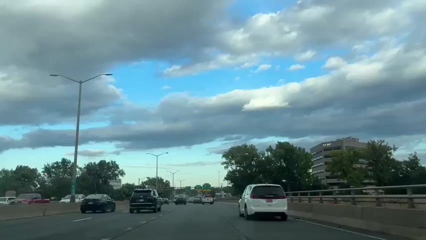 The video shows a point-of-view shot from a vehicle driving on a highway. The sky is partly cloudy with patches of blue visible. On the highway, there are other vehicles, including cars and a white minivan. Streetlights and trees line the sides of the road. In the distance, there is a multistory building with signage. The scene appears to be daytime.