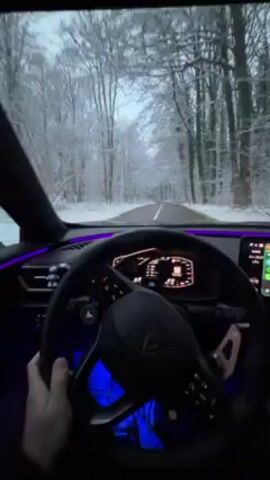 A first-person perspective video shows a driver behind the wheel of a car, driving on a road through a snowy forest. The interior of the car is visible, showcasing a modern dashboard with digital displays and ambient lighting, blue under the steering wheel and pink along the upper dash. The driver's hands are on the steering wheel, ready to use paddle shifters. The forest is covered in snow, with bare trees lining both sides of the road. The sky is overcast. The road is paved and appears to be relatively clear of snow, though snow is visible on the shoulder of the road.