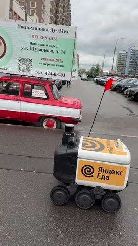 The video shows a white and yellow Yandex Eats delivery robot traversing a wet, paved area. The robot, which features six wheels, navigates past parked cars, puddles, and pedestrians. The background includes various shops and buildings, such as a pharmacy and a fitness center. Notably, a red and white car with a large advertisement sign mounted on its roof is parked along the street.