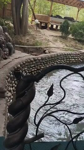The video shows a body of water, likely a stream or small river, flowing under a decorative metal railing. The railing is black and has a vine-like design with leaves. On the left side, there is a stone embankment with a pipe emerging from it, possibly a water feature. In the background, there is a wooden structure, resembling a gazebo or covered seating area, with tables and benches. Trees and vegetation are visible around the area, suggesting an outdoor, natural setting. The water appears somewhat murky, and the sunlight filters through the trees. There are no people visible in the video, and no explicit actions or interactions are taking place.