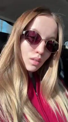 The video shows a woman with long, light-brown hair, wearing sunglasses with reddish-purple lenses and a red jacket. She is inside a vehicle and looks directly at the camera. She adjusts her sunglasses. Sunlight streams into the vehicle. The background is slightly blurred. Sounds of music are audible.