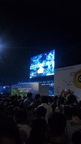 A large screen displays a performance on a stage, featuring a group of people wearing white and blue clothing. One person in the center is holding a reflective, multi-tiered object. The screen is set up outdoors at night, and a crowd of people is watching the performance. To the right of the screen, a sign is visible with Vietnamese text. The environment is dark.