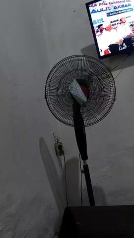 The video shows a standing fan in front of a light-colored wall. The fan has a circular metal grill and a black stand. A piece of green and white material is attached to the center of the fan grill. To the right of the fan, a television screen is visible, displaying images of people in white attire, possibly at an event. Text on the screen appears to be in a foreign language. An electrical outlet with a cord plugged into it is located on the wall behind the fan. The lighting in the room is dim, casting shadows on the wall.