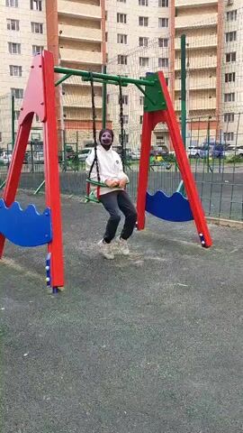 A person is swinging on a playground swing set. The person is wearing a white hoodie, dark pants, and light-colored sneakers. They have a dark mask covering their face, with only their eyes and forehead visible. The swing set has red A-frame legs with blue decorative elements and a green crossbar. The ground beneath the swing set is covered in a dark, soft material, likely rubberized safety surfacing.

In the background, a multi-story apartment building with many windows is visible. A chain-link fence separates the playground area from a parking lot where cars are parked. The lighting suggests it is daytime, and the weather appears to be overcast or cloudy, as there are no strong shadows. The scene is set in an urban or suburban residential area, consistent with the provided location context of Shchyolkovo, Russia. The person on the swing appears to be enjoying themselves, their body language relaxed as they move back and forth.