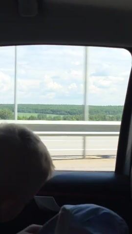 This is a video taken from inside a moving car, looking out the rear passenger window. In the foreground, the back of a person's head, with light-colored hair and wearing a grey shirt, is visible, slumped slightly forward. The person's head is positioned on the right side of the frame, partially obscuring the view out the window. The car's interior is dark, with a dark grey door panel and a silver door handle visible. A portion of a red trim is also visible on the door panel. Outside the window, a highway bridge is visible with white railings and road markings. Beyond the bridge, there is a vast landscape of green trees and fields under a bright blue sky with scattered white clouds. The video captures a sense of travel and observing the scenery from a vehicle.
