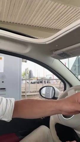 The video shows the interior of a car. A person's hands are visible in the foreground, holding a ticket. The steering wheel is in view, as is a smartphone mounted on the dashboard displaying a navigation app. Through the windshield, a highway and other vehicles can be seen. The scene appears to be at a toll booth or a similar checkpoint. The lighting suggests it is daytime.