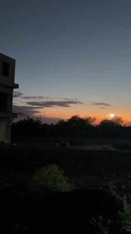 The video shows a rural landscape at dusk. A construction site with a concrete structure is on the left. The sky is a gradient of blue and orange, with a single star visible. There are trees and bushes in the middle ground, and a dirt field in the foreground. A streetlamp illuminates the scene from the right.