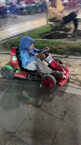 A young person wearing a blue hijab and a grey long-sleeved shirt is driving a red go-kart with the number 01 on the front. The go-kart has green lights on the back. The person's feet are resting on the pedals, and their hands are on the steering wheel. The go-kart is moving on a paved path. In the background, there are several people sitting at tables and chairs, with some tents and trees visible. The overall scene appears to be an outdoor recreational area or an amusement park at night, suggested by the artificial lighting.