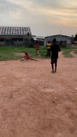 The video shows an outdoor scene with four people and a ball. The ground is made of dirt and sand. In the background, there are buildings that appear to be houses. The sky is visible, suggesting it is daytime, possibly late afternoon due to the lighting.

In the foreground, a person wearing a red shirt and red shorts is sitting on the ground with a ball. They appear to be holding the ball. To their left, another person is also sitting on the ground.

Further back, three other people are standing. One person is wearing a yellow shirt and dark shorts. Another person is wearing a black shirt and dark shorts, and they are walking away from the camera. The third standing person is wearing a patterned shirt and dark shorts.

The people appear to be engaged in some sort of activity, possibly a game of soccer, given the presence of the ball. The overall atmosphere seems casual and informal.