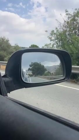 The video shows a view from inside a car, looking out the side window at the side mirror and the road ahead. The side mirror reflects the road behind the car, with other vehicles and trees visible. The car appears to be driving on a paved road in a rural area, with hills and vegetation on either side. The sky is partly cloudy. The video is a short clip, with no discernible sound.