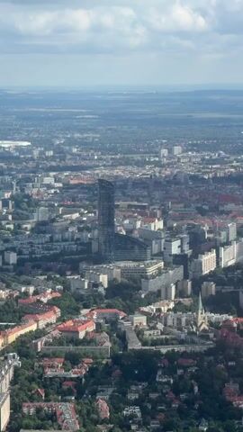 This is a vertical, high-angle aerial shot of a cityscape. In the center of the frame, a tall, modern skyscraper with a distinctive curved design stands out against a backdrop of lower-rise buildings. The surrounding city is dense, with a mix of residential and commercial structures, many featuring red-tiled roofs. Trees are interspersed throughout the urban landscape, particularly in the foreground and mid-ground. The sky above is partly cloudy. The overall impression is that of a sprawling, developed urban area.