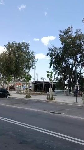 The video captures a street scene with a road in the foreground and a pedestrian area behind it. Several trees with green foliage line the pedestrian area, some of which are swaying in the wind. A building with a dark roof and glass walls is visible in the mid-ground, possibly a cafe or restaurant. A black car is parked on the left side of the street. The sky is blue with scattered white clouds. The overall impression is a calm, outdoor environment.