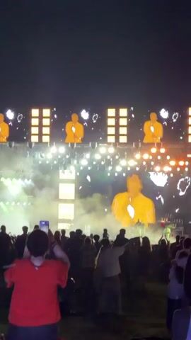 A concert is happening at night. A crowd of people are gathered in front of a large stage. There are many lights and screens on the stage, which display large images of a man. Some people in the crowd are holding up their phones to take pictures or videos. The person in the foreground is wearing a red shirt and is holding up a phone. The ground is covered in grass. There is smoke or fog in the air on the stage.