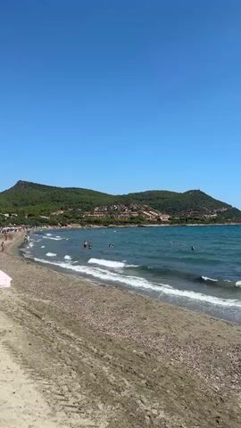 The video shows a crowded beach with people swimming in the ocean. The beach is sandy with some debris. The ocean is blue and has waves. In the background, there are green hills with some buildings on them and a clear blue sky.