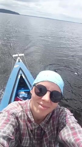 A person wearing sunglasses and a bandana is on a boat, holding a camera and filming themselves. The boat is moving through the water, creating a wake. The water is dark and choppy, and the sky is cloudy. In the background, there is a distant shoreline with trees. The person smiles and looks at the camera. The video is shot from a first-person perspective.