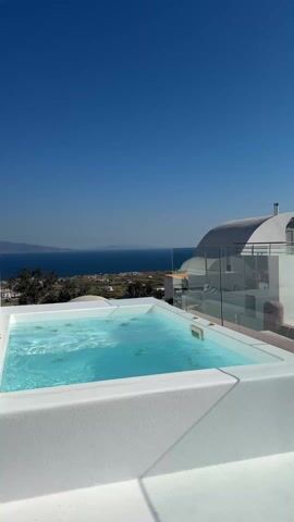 The video shows a panoramic view of Santorini, Greece, on a bright, sunny day. The video starts with a close-up of a small, white jacuzzi filled with clear water, overlooking the Aegean Sea. The camera pans to the right, revealing the typical white buildings of Santorini, with their unique architectural style. The buildings cascade down the hills, creating a picturesque landscape. A few people can be seen walking on the pathways among the buildings. In the background, the deep blue sea stretches towards the horizon, with a few boats visible. The sky is clear and blue, without any clouds. The overall atmosphere is serene and picturesque.