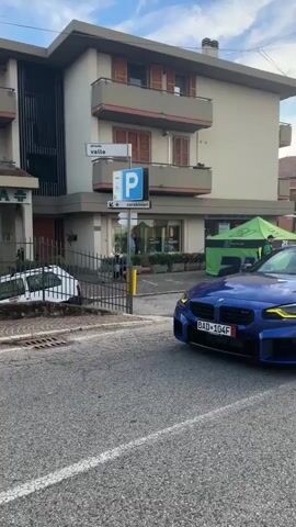 A blue BMW M2 drives down a street. The car has tinted windows and black wheels. The driver is visible in the driver's seat. In the background, there is a building with balconies and shutters. A sign with a blue "P" indicates parking. A white car is parked on the left side of the street. A second car, a white BMW with racing decals, drives past in the opposite direction. The street is paved and has white lane markings. The sky is cloudy.