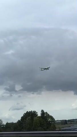 An airplane, painted white with a green tail and accents, descends through a cloudy sky towards a runway. Below the plane, the landscape features trees, a large solar panel field, and buildings in the distance. The view is captured from a moving vehicle, as evidenced by the blurred foreground of a guardrail and road. The sky is overcast, and the overall atmosphere is grey.
